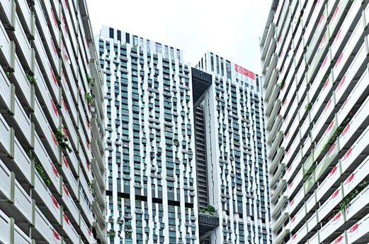 A modern urban architectural scene features high-rise buildings with a distinctive grid-like pattern. The skyscrapers have a mix of vertical and horizontal lines, creating an intricate façade. Singapore flags are displayed on the buildings, and greenery is integrated into the structure, reflecting urban planning and environmental considerations.
