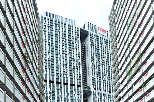 A modern urban architectural scene features high-rise buildings with a distinctive grid-like pattern. The skyscrapers have a mix of vertical and horizontal lines, creating an intricate façade. Singapore flags are displayed on the buildings, and greenery is integrated into the structure, reflecting urban planning and environmental considerations.