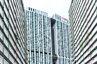 A modern urban architectural scene features high-rise buildings with a distinctive grid-like pattern. The skyscrapers have a mix of vertical and horizontal lines, creating an intricate fa&ccedil;ade. Singapore flags are displayed on the buildings, and greenery is integrated into the structure, reflecting urban planning and environmental considerations.