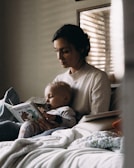 A woman is sitting on a bed, holding a child in her lap. The child is dressed in pajamas and is holding a book titled 'Tumble Bumble' while sucking on a pacifier. The setting appears to be cozy, with soft lighting filtering through a window blind, creating a calm and intimate atmosphere.
