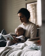 A woman is sitting on a bed, holding a child in her lap. The child is dressed in pajamas and is holding a book titled 'Tumble Bumble' while sucking on a pacifier. The setting appears to be cozy, with soft lighting filtering through a window blind, creating a calm and intimate atmosphere.