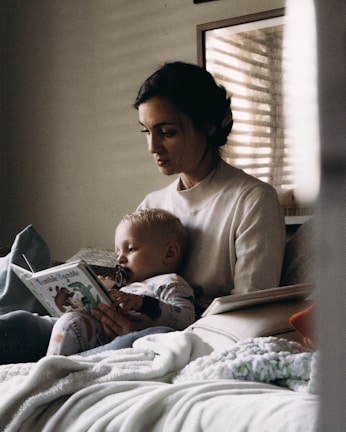 A woman is sitting on a bed, holding a child in her lap. The child is dressed in pajamas and is holding a book titled 'Tumble Bumble' while sucking on a pacifier. The setting appears to be cozy, with soft lighting filtering through a window blind, creating a calm and intimate atmosphere.