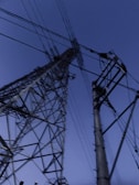 Large electrical transmission towers stretch upwards against a deep blue sky. The towers are composed of intricate metal frameworks and numerous power lines crisscross the scene.