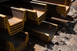 Stacks of large steel beams with yellow edges are arranged on a muddy construction site. The beams are layered in a repeating pattern and there are scattered rocks and debris on the ground.