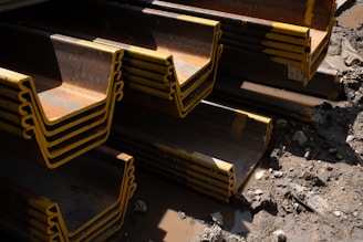 Stacks of large steel beams with yellow edges are arranged on a muddy construction site. The beams are layered in a repeating pattern and there are scattered rocks and debris on the ground.