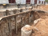 A construction site featuring a large excavation with exposed soil and rocky layers. Wooden supports and barriers are used to secure the sides of the pit. In the background, temporary structures or containers are visible, along with safety barriers.