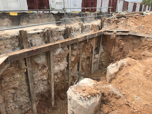 A construction site featuring a large excavation with exposed soil and rocky layers. Wooden supports and barriers are used to secure the sides of the pit. In the background, temporary structures or containers are visible, along with safety barriers.
