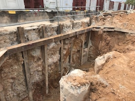 A construction site featuring a large excavation with exposed soil and rocky layers. Wooden supports and barriers are used to secure the sides of the pit. In the background, temporary structures or containers are visible, along with safety barriers.