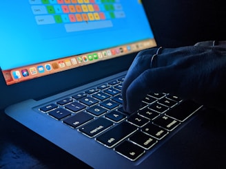 A hand is typing on a dimly lit laptop keyboard with an illuminated screen displaying a colorful keyboard layout. The environment appears dark, emphasizing the glow from the keys and screen.