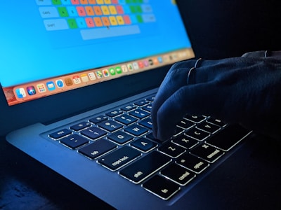 Close-up of a person’s hands typing on a laptop keyboard in dim light, with green code reflections on the screen.