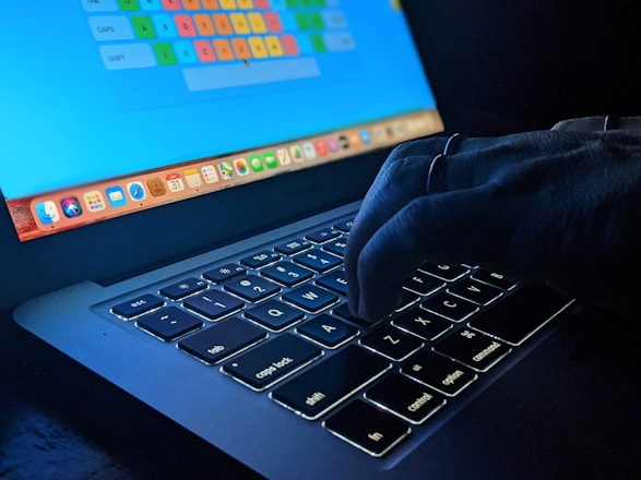 A hand is typing on a dimly lit laptop keyboard with an illuminated screen displaying a colorful keyboard layout. The environment appears dark, emphasizing the glow from the keys and screen.