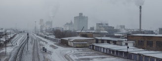 An industrial area with a railway track runs parallel to several warehouse buildings. Smoke billows from chimneys in the background, contributing to a grey, overcast sky. The ground is lightly dusted with snow, indicating cold weather conditions.