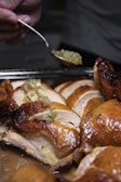 Sliced roasted chicken with golden-brown skin is arranged on a tray. A hand holding a spoon is adding a green onion and oil mixture on top of the chicken, adding a glistening effect. The texture of the chicken is moist and juicy.