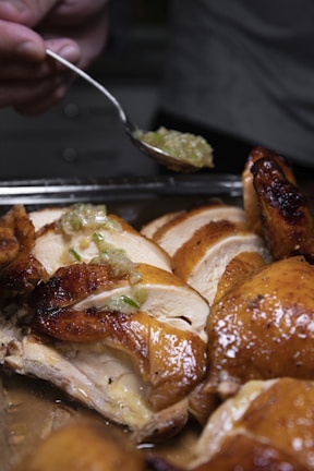 Sliced roasted chicken with golden-brown skin is arranged on a tray. A hand holding a spoon is adding a green onion and oil mixture on top of the chicken, adding a glistening effect. The texture of the chicken is moist and juicy.