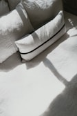An arrangement of pillows placed on a bed, with sunlight casting shadows over them. The pillows appear to be in different shapes and sizes, with the prominent one being rectangular and featuring a dark stripe design.