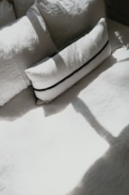 An arrangement of pillows placed on a bed, with sunlight casting shadows over them. The pillows appear to be in different shapes and sizes, with the prominent one being rectangular and featuring a dark stripe design.