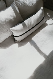 An arrangement of pillows placed on a bed, with sunlight casting shadows over them. The pillows appear to be in different shapes and sizes, with the prominent one being rectangular and featuring a dark stripe design.