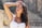 A woman with long, flowing dark hair stands with her eyes closed, head tilted slightly back and one hand resting above her forehead. She wears a white sleeveless top with button details. The background is a blurred outdoor setting suggesting a sunny day.