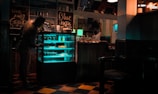 A dimly lit cafe interior with a person leaning towards a refrigerated display case filled with illuminated shelves. The decor includes handwritten chalkboard signs with phrases like 'Take Away Gelato' and 'Blue Kejir'. Another person is seen in the background reaching for something on a shelf. The floor has a checkered pattern, and there are various items and bottles on the counter.