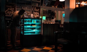 A dimly lit cafe interior with a person leaning towards a refrigerated display case filled with illuminated shelves. The decor includes handwritten chalkboard signs with phrases like 'Take Away Gelato' and 'Blue Kejir'. Another person is seen in the background reaching for something on a shelf. The floor has a checkered pattern, and there are various items and bottles on the counter.