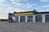 A modern industrial building with a corrugated metal facade featuring large garage doors and a yellow accent stripe. A white semi-truck is parked in front of the building. The sky is partly cloudy, and the surrounding area is an open gravel lot.