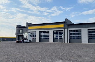 A modern industrial building with a corrugated metal facade featuring large garage doors and a yellow accent stripe. A white semi-truck is parked in front of the building. The sky is partly cloudy, and the surrounding area is an open gravel lot.