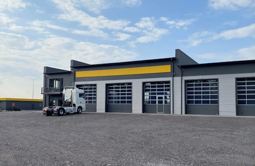 A modern industrial building with a corrugated metal facade featuring large garage doors and a yellow accent stripe. A white semi-truck is parked in front of the building. The sky is partly cloudy, and the surrounding area is an open gravel lot.