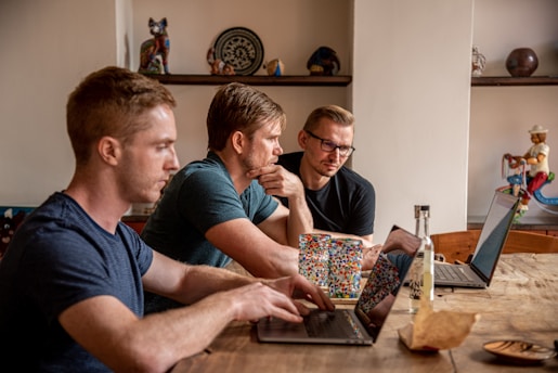 Three men are seated at a wooden table, each working on a laptop. The setting appears to be a cozy room with decorative items on shelves in the background, including a colorful mug and a figurine. One man is focused on his laptop, while the other two are engaging in a discussion, possibly collaborating on a work project.