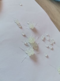 A cluster of small, translucent adhesive glue drops or beads arranged on a white sheet of paper, with delicate and thin strings connecting some of them. The surface of the paper is slightly textured and the lighting creates subtle shadows.