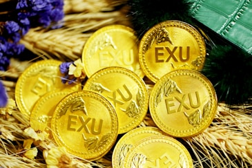 A collection of gold coins with the inscription 'EXU' is placed on a bed of wheat stalks. Purple and yellow flowers are scattered among the coins. A green, textured object, possibly a wallet or book, is partially visible at the top right.