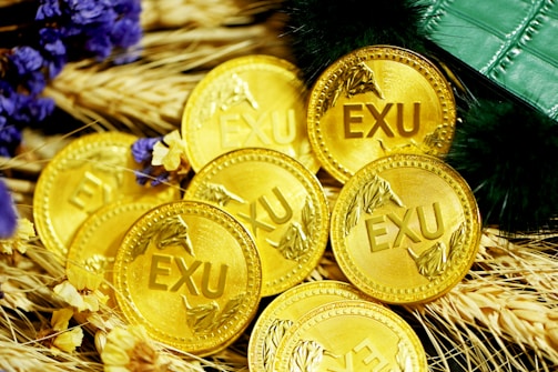 A collection of gold coins with the inscription 'EXU' is placed on a bed of wheat stalks. Purple and yellow flowers are scattered among the coins. A green, textured object, possibly a wallet or book, is partially visible at the top right.