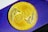 A gold coin featuring embossed lettering 'EXU' with a decorative design reminiscent of wings or feathers on either side. The background consists of a purple surface with subtle geometric patterns.
