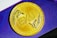 A gold coin featuring embossed lettering 'EXU' with a decorative design reminiscent of wings or feathers on either side. The background consists of a purple surface with subtle geometric patterns.