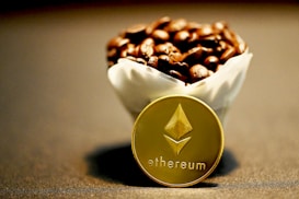 A close-up view of a cup filled with coffee beans, with a golden Ethereum coin placed in front. The background is blurred, creating a focus on the coin and the beans.