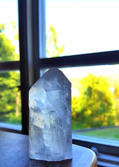 A transparent crystal with sharp edges sits on a wooden table near a window with a bright view of lush green trees. Sunlight illuminates the crystal, casting subtle reflections and highlighting intricate details.
