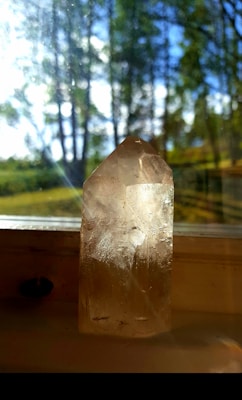 A clear, faceted crystal stands on a windowsill with sunlight streaming through the glass, creating reflections and a play of light. In the background, blurry green trees and a clear blue sky suggest an outdoor setting.