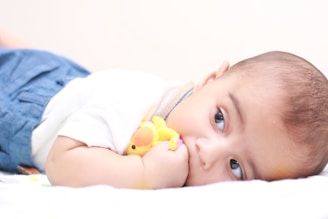A baby is lying on its stomach, gazing curiously to the side. The baby is holding a small yellow plush toy close to its mouth. The baby is wearing a light-colored top and blue shorts, and is resting on a white surface.