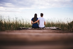 man and woman sitting on brown wooden pathway surrounded by green grass during daytime