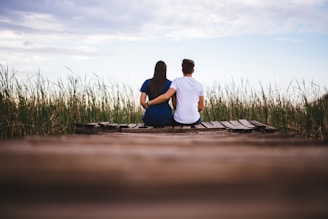 man and woman sitting on brown wooden pathway surrounded by green grass during daytime