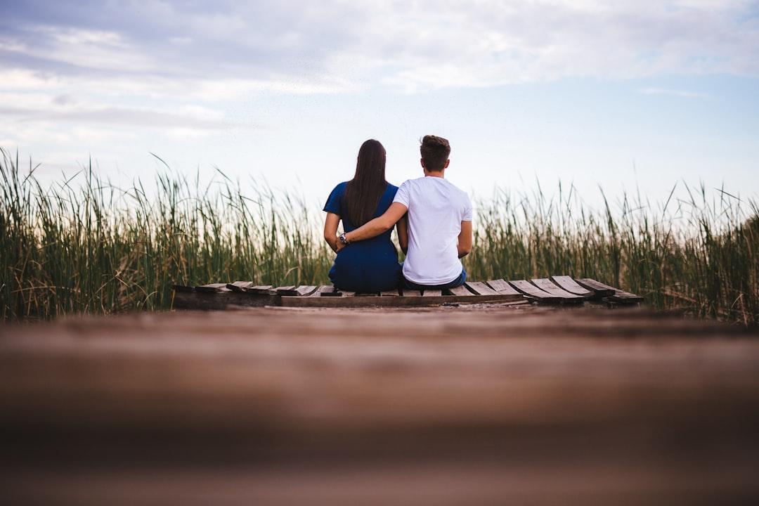 man and woman sitting on brown wooden pathway surrounded by green grass during daytime