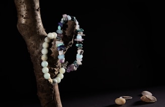 A wooden branch positioned against a dark background with two bead necklaces draped over it. One necklace is made of rounded turquoise beads, while the other features irregularly shaped stones in various shades of blue and purple. On a dark surface below, a few smooth pebbles and a single silver ring are visible.