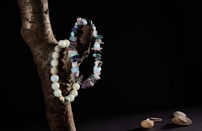 A wooden branch positioned against a dark background with two bead necklaces draped over it. One necklace is made of rounded turquoise beads, while the other features irregularly shaped stones in various shades of blue and purple. On a dark surface below, a few smooth pebbles and a single silver ring are visible.