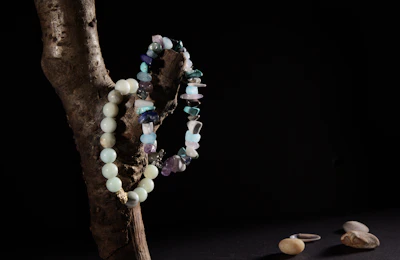 A wooden branch positioned against a dark background with two bead necklaces draped over it. One necklace is made of rounded turquoise beads, while the other features irregularly shaped stones in various shades of blue and purple. On a dark surface below, a few smooth pebbles and a single silver ring are visible.