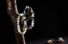 A wooden branch positioned against a dark background with two bead necklaces draped over it. One necklace is made of rounded turquoise beads, while the other features irregularly shaped stones in various shades of blue and purple. On a dark surface below, a few smooth pebbles and a single silver ring are visible.
