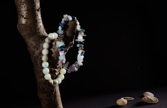 A wooden branch positioned against a dark background with two bead necklaces draped over it. One necklace is made of rounded turquoise beads, while the other features irregularly shaped stones in various shades of blue and purple. On a dark surface below, a few smooth pebbles and a single silver ring are visible.