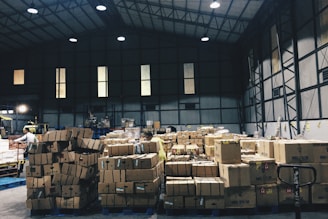 A warehouse interior filled with stacks of cardboard boxes arranged on pallets. The space is large and industrial with high ceilings and metal beams. There is a worker seen handling the boxes.