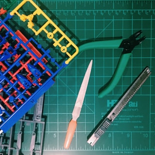 A collection of hobby tools and plastic model parts arranged on a green cutting mat with a grid pattern. The items include plastic model sprues in blue, red, and yellow, a metal file with an orange handle, green-handled clippers, and a utility knife.