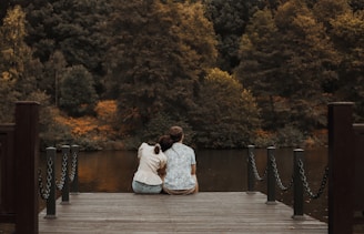 A couple sits on a wooden dock facing a tranquil lake surrounded by dense forest. The foliage shows a mix of green and hints of autumn colors. They are seated closely, creating a serene and intimate atmosphere.
