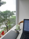 A modern workspace setup by a bright window featuring a lush green view outside with leafy trees. On the desk, there's a glass of iced green drink with a straw, a closed black notebook, and an open laptop displaying file windows. The Vietnamese flag is visible outside, adding a touch of national identity.