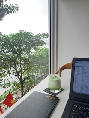 A modern workspace setup by a bright window featuring a lush green view outside with leafy trees. On the desk, there's a glass of iced green drink with a straw, a closed black notebook, and an open laptop displaying file windows. The Vietnamese flag is visible outside, adding a touch of national identity.